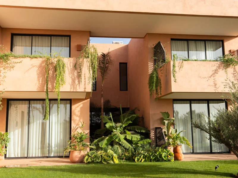 Terracotta-rendered villa facade at Dar Manou in Marrakech, with dark lattice screens, planted terraces spilling greenery over the parapets, and a palm and olive tree on the lawn in late afternoon sun