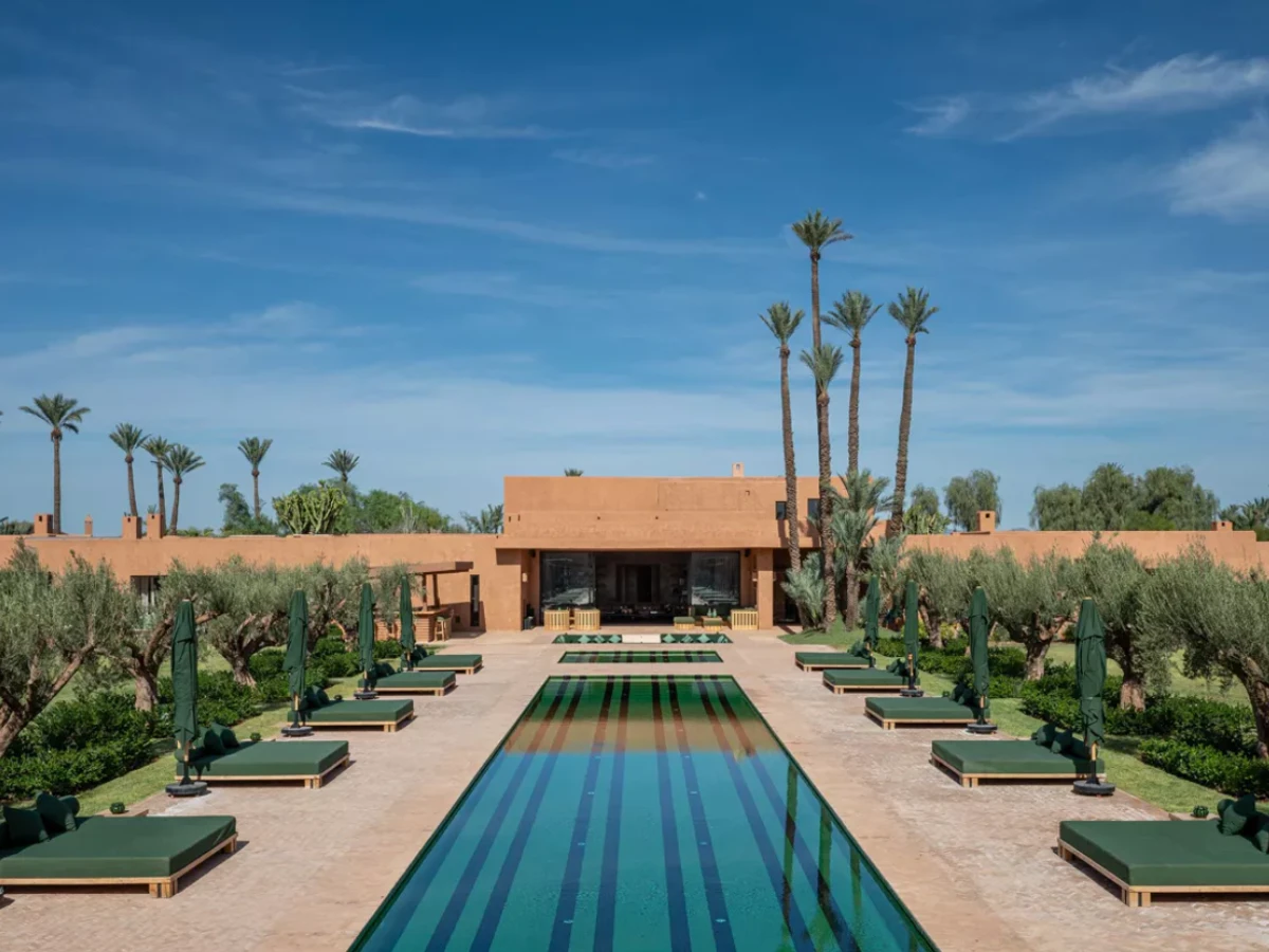 The pool terrace at Villa Kemgia outside Marrakech, paired sun loungers and parasols along a long rectangular pool, clipped hedges, olive trees and date palms beyond the terracotta boundary wall