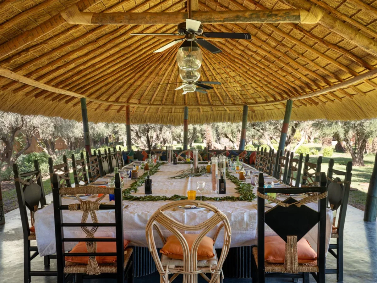 A long dining table set for a group under a thatched pavilion among olive trees at Villa Kemgia near Marrakech